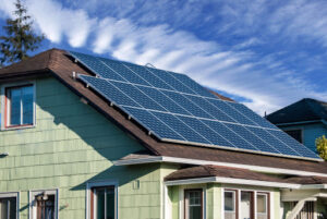 A residential home is pictured with solar panels installed on the roof. The home is light green with a brown roof. It looks to be built during the mid-20th century.